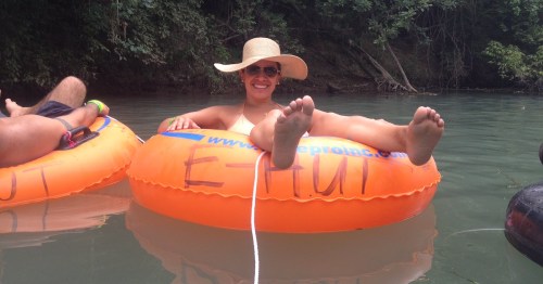 A woman relaxes on an orange inner tube in a river, wearing a wide-brimmed hat and smiling at the camera.