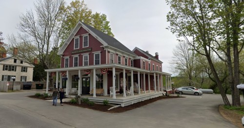A red historic building with a porch, surrounded by trees, and two people standing outside on a cloudy day.