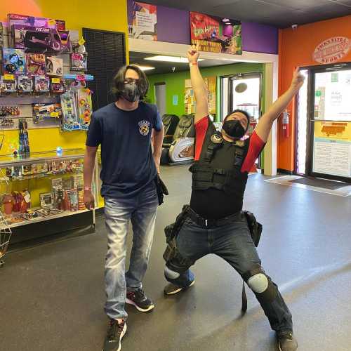 Two people in a colorful store; one is celebrating with arms raised, while the other stands nearby, both wearing masks.