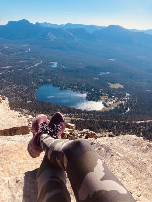 A person sits on a rocky ledge, legs crossed, overlooking a scenic mountain landscape and a shimmering lake below.
