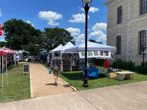 A man walks along a path at an outdoor market with tents and a historic building in the background under a blue sky.