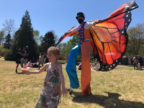 A child joyfully runs while a stilt performer in a butterfly costume poses nearby on a sunny day in a park.