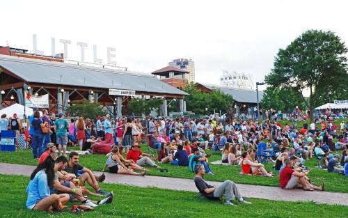 A large crowd gathers on a grassy area at an outdoor event, with buildings and a sign reading "LITTLE" in the background.