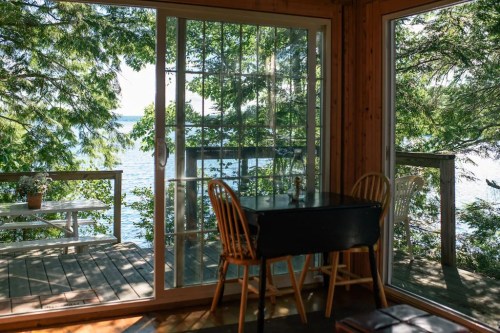 A cozy interior with a table and chairs, overlooking a serene lake through large glass doors surrounded by trees.