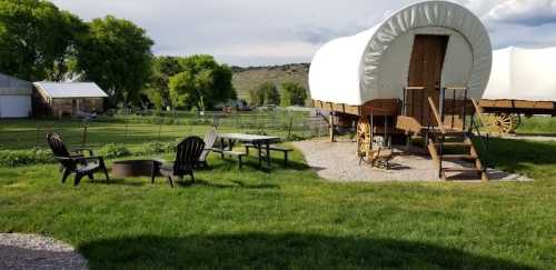 A grassy area with wooden chairs and a fire pit, featuring two covered wagons in the background.