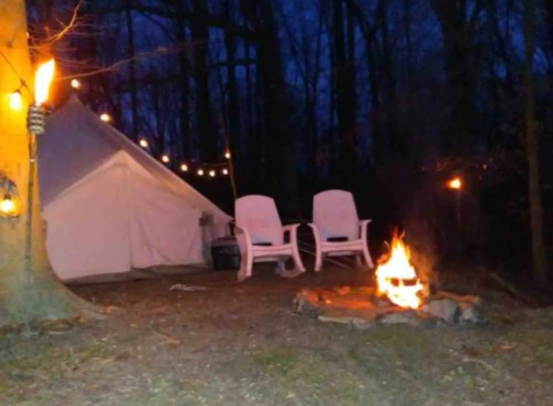 A cozy campsite at night with a fire pit, two chairs, and a tent surrounded by trees and string lights.