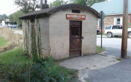 Small, weathered building labeled "Old Flippin City Jail, circa 1933," surrounded by grass and parked cars.