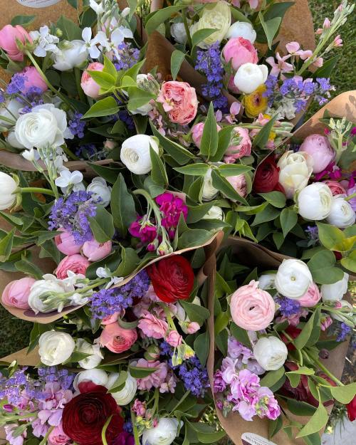 A vibrant assortment of colorful flowers in brown paper wraps, featuring roses, ranunculus, and various greenery.