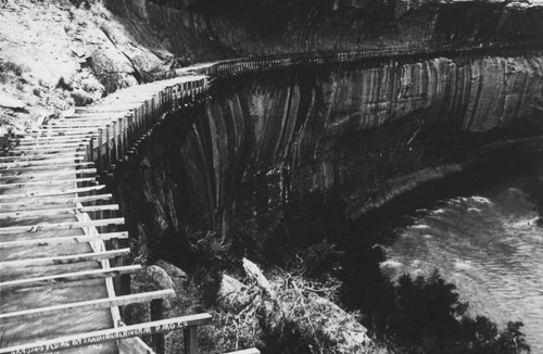 A narrow wooden walkway along a steep cliff overlooking a river, surrounded by rugged rock formations.