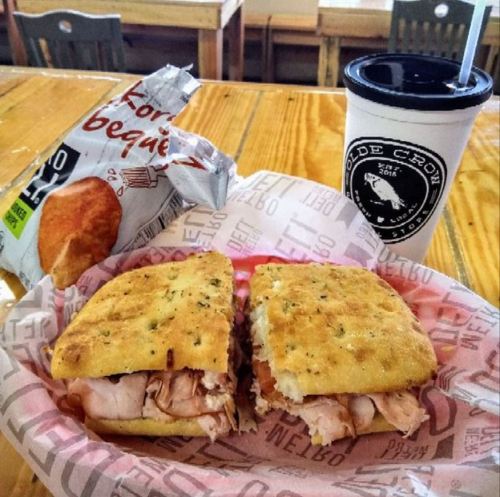 A sandwich with turkey on a plate, a bag of chips, and a drink in a cup on a wooden table.