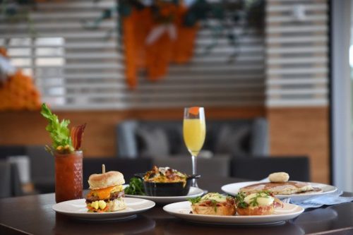 A table with a variety of dishes: a burger, eggs benedict, a skillet, and a mimosa, set in a cozy dining area.