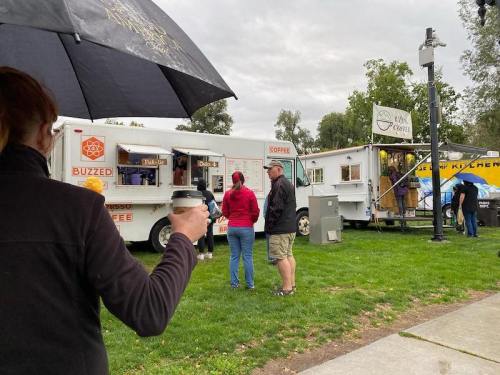 A person holding a coffee cup stands under an umbrella, watching people at food trucks in a park on a cloudy day.