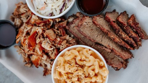 A platter featuring sliced brisket, pulled pork, coleslaw, and macaroni and cheese in bowls with barbecue sauce.