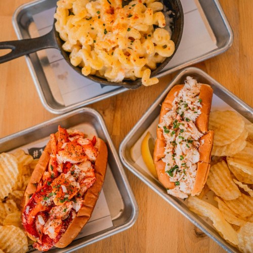 A tray with two lobster rolls, a bowl of mac and cheese, and a side of potato chips on a wooden table.