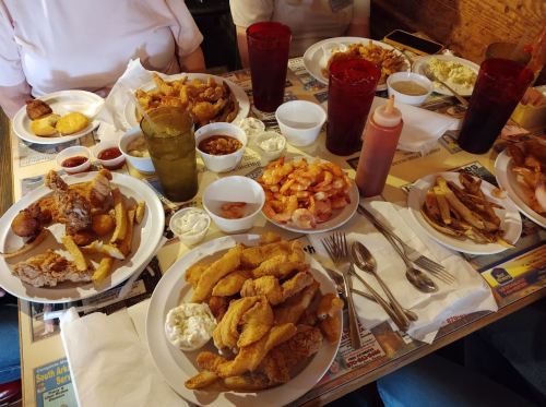 A table filled with various plates of fried seafood, sides, sauces, and drinks in a casual dining setting.