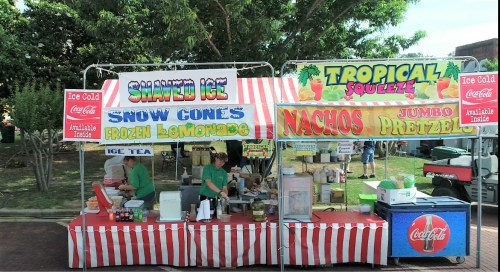 A colorful food stand featuring shaved ice, snow cones, nachos, and jumbo pretzels, with two vendors in green shirts.