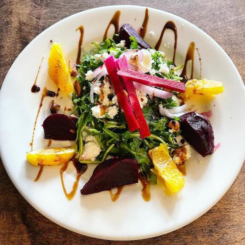 A vibrant salad with arugula, beets, orange slices, goat cheese, and drizzled balsamic glaze on a white plate.