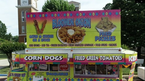 Food stand featuring a colorful sign with "Bloomin' Onion," "Fried Pickles," and various snacks like corn dogs and fried green tomatoes.