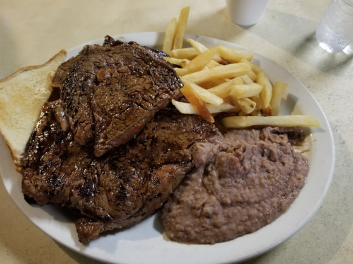 A plate of grilled steak, French fries, refried beans, and a slice of bread on a table.