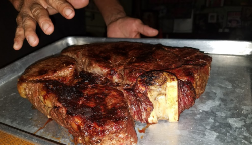 A large, well-cooked steak on a metal tray, with a hand reaching towards it in the background.