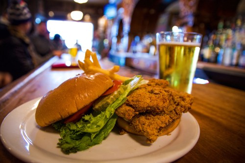 A plate with a fried chicken sandwich, lettuce, tomato, and fries, alongside a glass of beer at a bar.