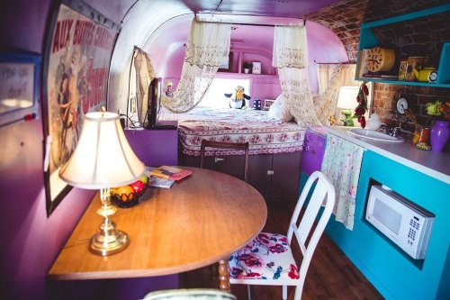 A cozy, colorful vintage trailer interior with a bed, dining table, and kitchenette, featuring floral decor and warm lighting.