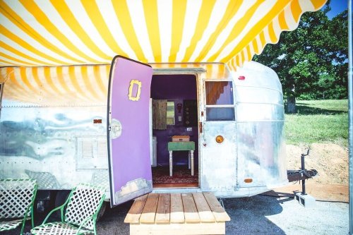 A colorful vintage trailer with a striped awning, open door, and outdoor seating area.
