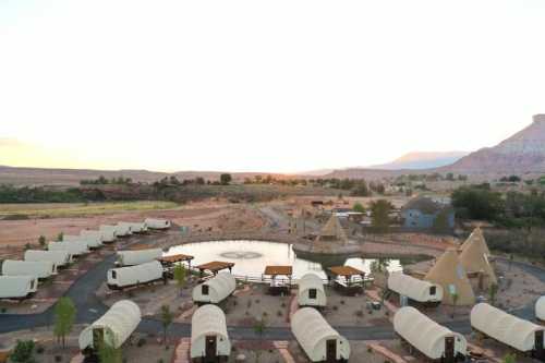 Aerial view of a glamping site with tents arranged around a pond, set against a sunset backdrop and rocky landscape.