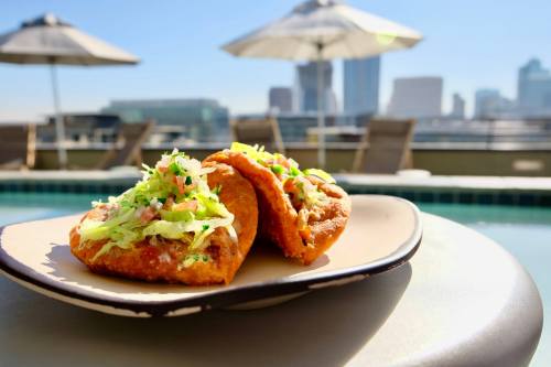 Two crispy tacos topped with lettuce and tomatoes, set against a sunny rooftop poolside backdrop.