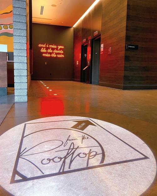 A stylish lobby with elevator doors and a circular logo on the floor reading "from the Rooftop." A quote is on the wall.