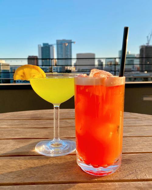 Two colorful cocktails on a wooden table with a city skyline in the background during sunset.