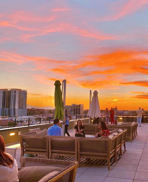 People enjoying a rooftop gathering at sunset, with vibrant orange and pink skies and city buildings in the background.