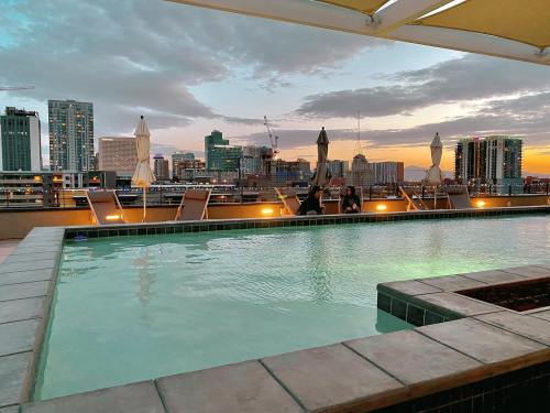 Rooftop pool at sunset with city skyline, lounge chairs, and umbrellas under a colorful sky.