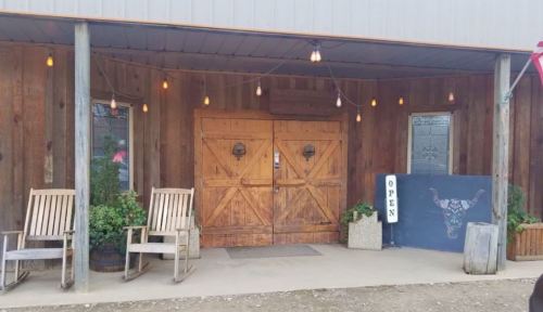 Rustic wooden entrance with double doors, rocking chairs, potted plants, and an "OPEN" sign under warm hanging lights.