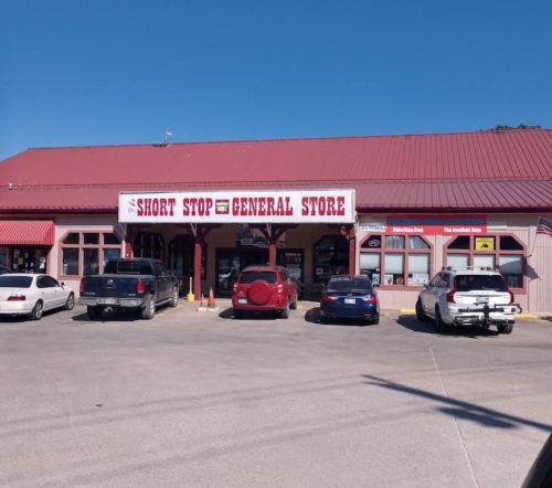 A general store with a red roof and sign, surrounded by parked cars on a sunny day.