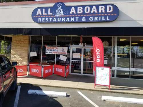 Exterior of "All Aboard Restaurant & Grill" with signs for delivery and curbside pickup, featuring red flags and a chalkboard.
