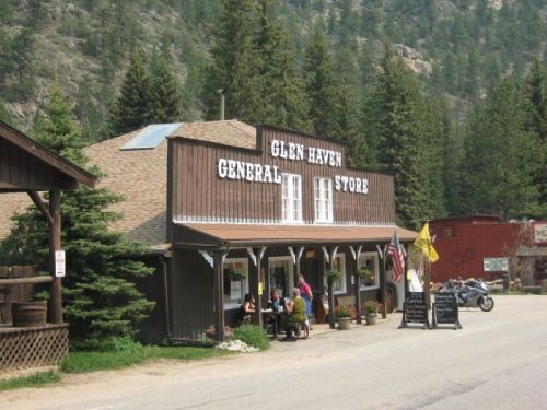 A rustic general store with a wooden facade, surrounded by trees, featuring outdoor seating and a motorcycle parked nearby.