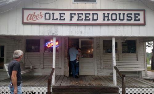 A rustic building with a sign reading "Abe's Ole Feed House" and an "Open" neon sign, featuring two people at the entrance.