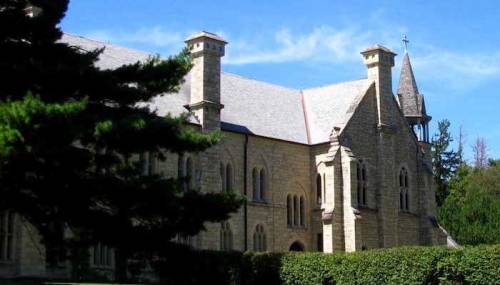 A historic stone building with pointed towers, surrounded by trees and blue sky.