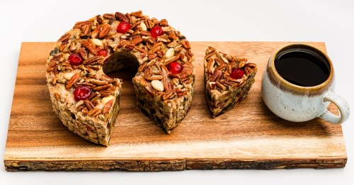 A round fruitcake topped with nuts and cherries, with a slice cut out, next to a cup of coffee on a wooden board.