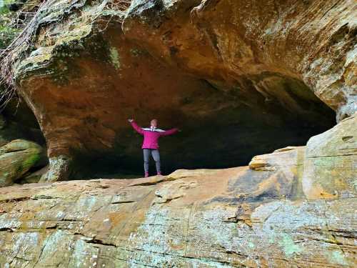 A person stands with arms outstretched on a rocky ledge inside a cave, surrounded by natural stone formations.