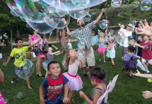 A lively outdoor scene with children playing and dancing among large bubbles, with a man joyfully engaging with them.