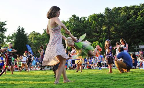 A woman twirls a young child in a grassy area, surrounded by children playing with hula hoops and enjoying a summer event.
