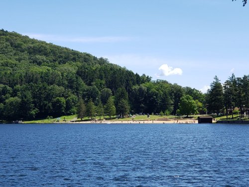 A serene lake surrounded by lush green hills, with a sandy beach and people enjoying the outdoors on a sunny day.