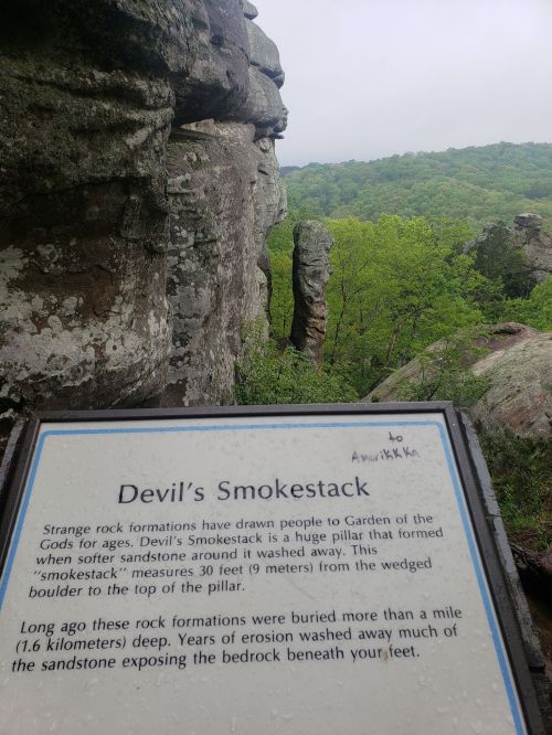 Sign about "Devil's Smokestack," a rock formation in Garden of the Gods, with lush green hills in the background.