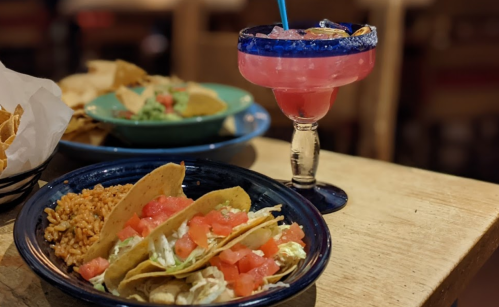 A plate of tacos with tomatoes and lettuce, served with rice, alongside a colorful cocktail in a glass.