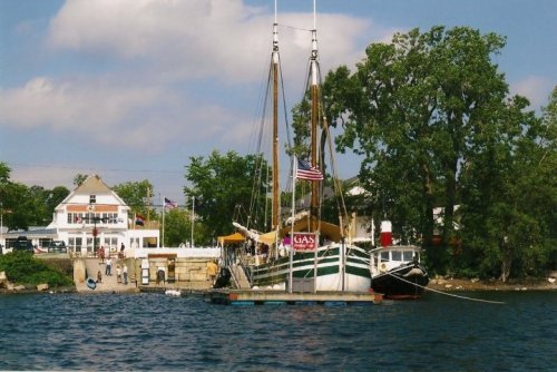 A dock with a tall ship and a smaller boat, surrounded by trees and a building, under a partly cloudy sky.
