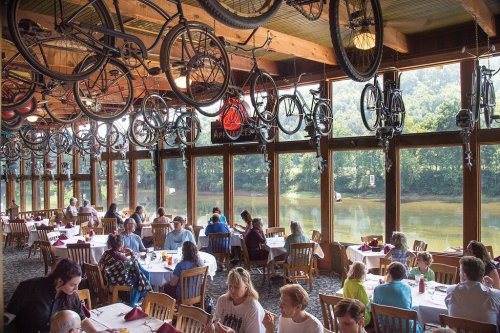 A bustling restaurant with bicycles hanging from the ceiling, overlooking a scenic river view through large windows.