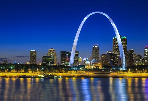 The Gateway Arch illuminated at night, reflecting in the water with a city skyline in the background.