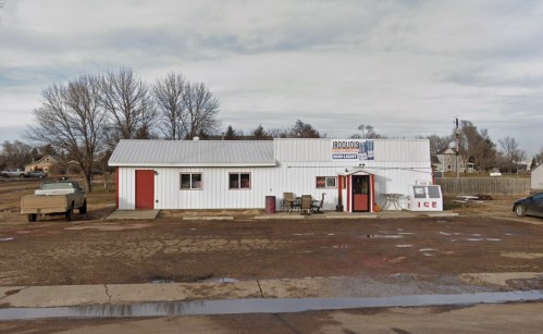 A small white building with a metal roof, featuring a sign for Bud Light and an ice sales window, surrounded by gravel.